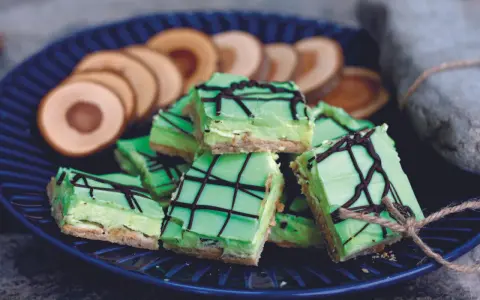 Gröna biskvier i långpanna med chokladmönster, serverade på en blå tallrik med några runda kex i bakgrunden.