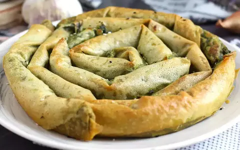 En gyllene, spiralformad burek med spenatfyllning, perfekt för ett hemlagat burek recept. Vitlök och lök i bakgrunden.