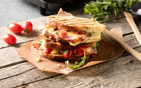 En staplad grilled cheese med skinka, tomater och ruccola på en träyta.