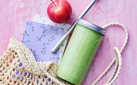En grön, proteinrik smoothie i glasburk med metallrör, bredvid ett rött äpple och en vävd kasse på en rosa bakgrund.