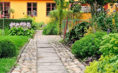 En stenlagd gång leder genom en lummig trädgård med en gul fasad. Bland gröna växter syns lila blommor och några små orangea blommor som lyser upp.