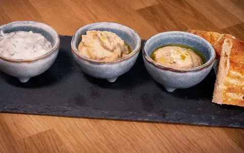 Tre skålar med kall mat, troligen dippsåser eller röror, serveras med rostat bröd på en svart stenplatta.