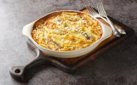 En krämig Janssons frestelse med potatis, ansjovis och ströbröd i en ugnsform, redo att serveras med två silvergafflar.