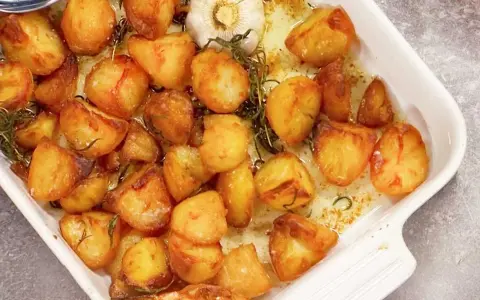 Ugnsrostad potatis i en vit form, gyllene och krispiga, med en hel vitlöksklyfta och rosmarin.