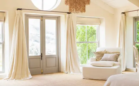 Ett rum med dubbeldörrar och fönster, draperade i ljusa gardiner som hänger från en tunn gardinstång. En vit fåtölj med kuddar står vid ett fönster.