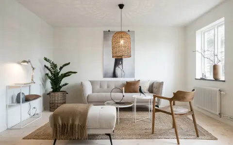 En ljus och luftig lounge med en bekväm soffa, en pall med en filt i honungsgyllene nyanser och en trästol. En stor flätad lampa ger ett varmt sken.