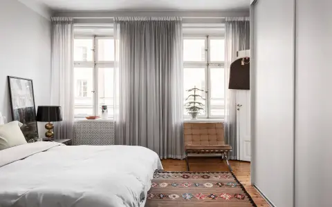 Sovrum med en stor säng, en läderfåtölj och ljusa gardiner som släpper in dagsljus. En matta med mönster ligger på trägolvet.