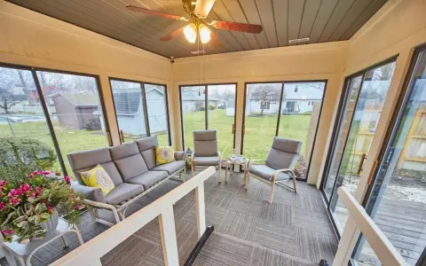 En inredd veranda med grå soffa, fåtöljer och ett lågt bord. Stora fönster ger utsikt över en grönskande trädgård.