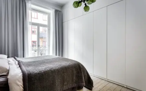 Sovrum med en stor, vit garderob med draperi. En säng med mörkt överkast står framför ett fönster med ljusgrå gardiner.
