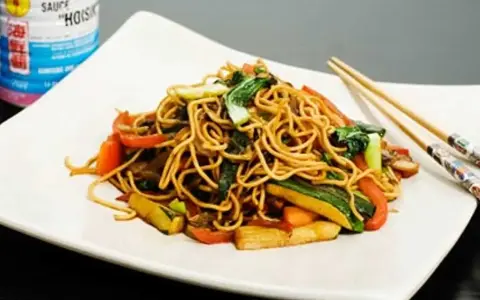 En tallrik med nudlar och grönsaker, perfekt för ett snabbt wok recept. Hoisinsåsburk syns i bakgrunden.