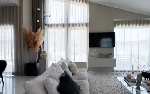 En ljus och luftig vardagsrumsmiljö med en stor soffa, ett soffbord och en TV. Varma gardiner släpper in ett mjukt ljus.