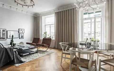 Vardagsrum med många fönster, draperade i ljusa gardiner. Två bruna läderfåtöljer och ett soffbord med böcker.