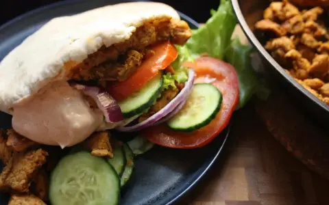 En saftig kyckling kebab i pitabröd med grönsaker och sås. Bredvid ligger mer kyckling i en panna.