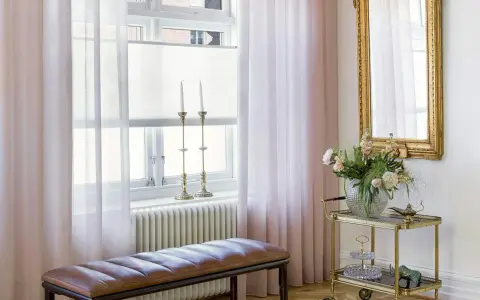 Ljusrosa gardiner längd från golvet ramar in ett fönster med ljusinsläpp. En brun läderbänk står framför ett element.