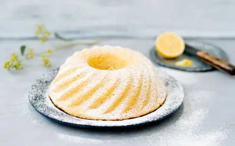 En vacker citronkaka i långpanna, pudrad med florsocker, står på ett fat. En halv citron och en kniv syns i bakgrunden.