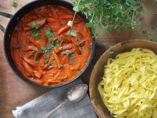 En krämig korv stroganoff med morötter i en panna, bredvid en skål med färsk pasta.