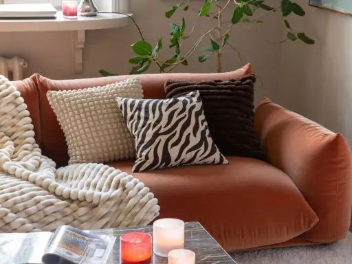 En orange soffa med flera kuddar och en mjuk pläd. Fina dekorationer som ljus och en tidning på soffbordet skapar en mysig atmosfär.