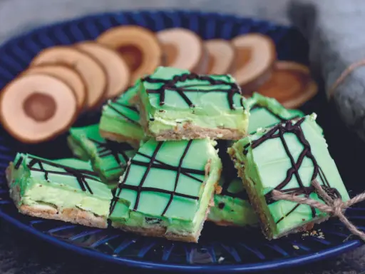 Gröna biskvier i långpanna med chokladmönster, serverade på en blå tallrik med några runda kex i bakgrunden.