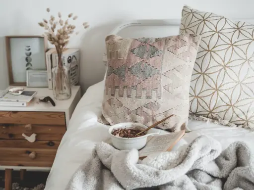 En säng med kuddar, en skål med frukost och en bok. Nattduksbordet har en vas med torkade blommor och en månkalender. En mysig och udda inredning.