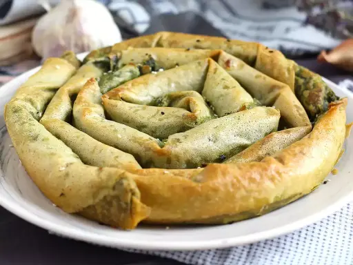 En gyllene, spiralformad burek med spenatfyllning, perfekt för ett hemlagat burek recept. Vitlök och lök i bakgrunden.