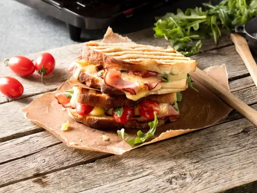 En staplad grilled cheese med skinka, tomater och ruccola på en träyta.
