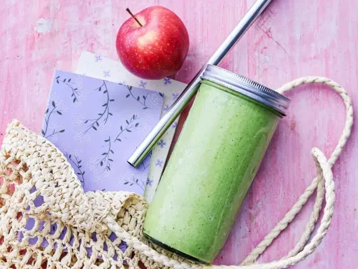 En grön, proteinrik smoothie i glasburk med metallrör, bredvid ett rött äpple och en vävd kasse på en rosa bakgrund.