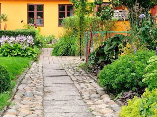 En stenlagd gång leder genom en lummig trädgård med en gul fasad. Bland gröna växter syns lila blommor och några små orangea blommor som lyser upp.