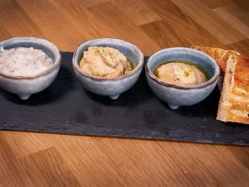 Tre skålar med kall mat, troligen dippsåser eller röror, serveras med rostat bröd på en svart stenplatta.