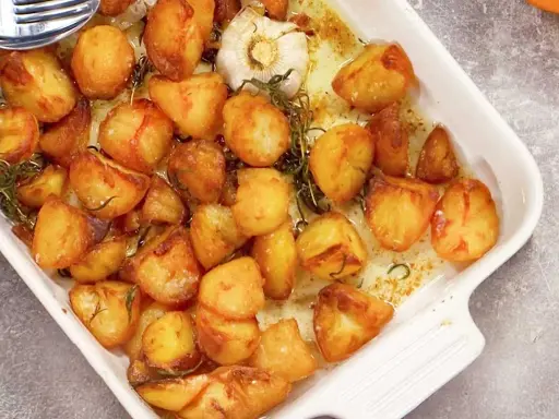 Ugnsrostad potatis i en vit form, gyllene och krispiga, med en hel vitlöksklyfta och rosmarin.