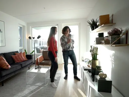 Två personer diskuterar något i ett modernt vardagsrum med trendig inredning, mörk soffa och hyllor med växter.