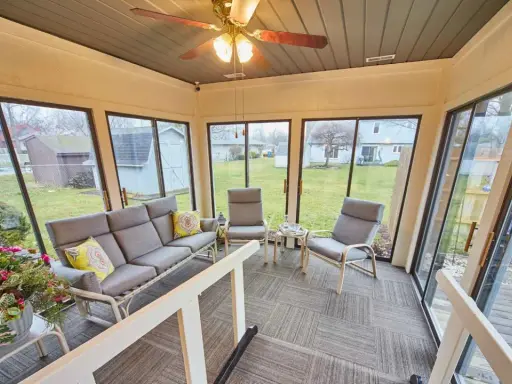 En inredd veranda med grå soffa, fåtöljer och ett lågt bord. Stora fönster ger utsikt över en grönskande trädgård.