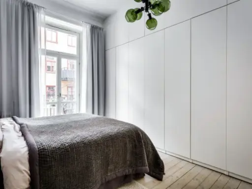 Sovrum med en stor, vit garderob med draperi. En säng med mörkt överkast står framför ett fönster med ljusgrå gardiner.