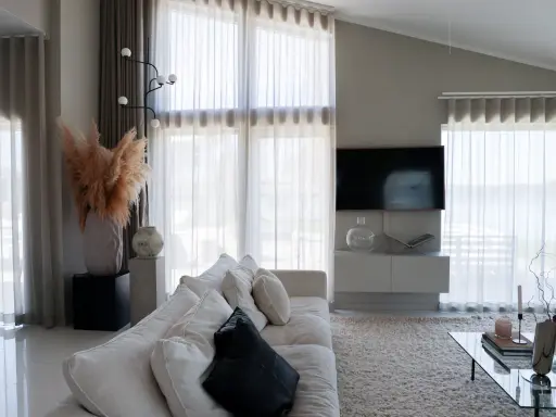 En ljus och luftig vardagsrumsmiljö med en stor soffa, ett soffbord och en TV. Varma gardiner släpper in ett mjukt ljus.