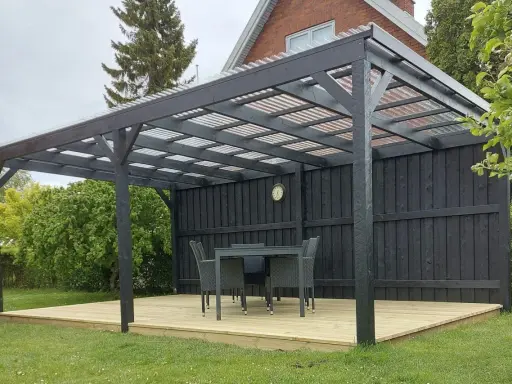 En mörk paviljong på en träaltan med bord och stolar. Perfekt för fika i trädgården.