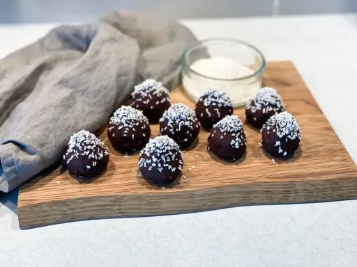 Mörka, chokladöverdragna protein chokladbollar toppade med kokosflingor ligger på en träskärbräda.