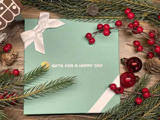 En grön presentask med texten "GIFTS FOR A HAPPY DAY", dekorerad med julgranspynt, kottar och röda bär. Perfekt för julklappar och julrim.