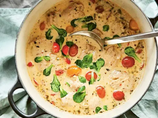 En krämig provencalsk kycklinggryta med körsbärstomater och gröna blad i en gjutjärnsgryta, redo att serveras med en slev.