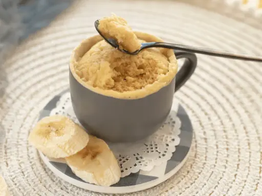 En fluffig mugcake proteinpulver med bananskivor. En sked lyfter en bit av den läckra kakan.