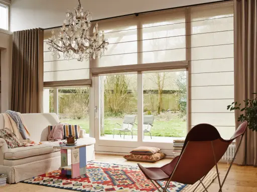 En ljus vardagsrumsmiljö med en bekväm soffa, en färgglad matta och en elegant kristallkrona. En hissgardin på gardinstång ger ett mjukt ljusinsläpp.