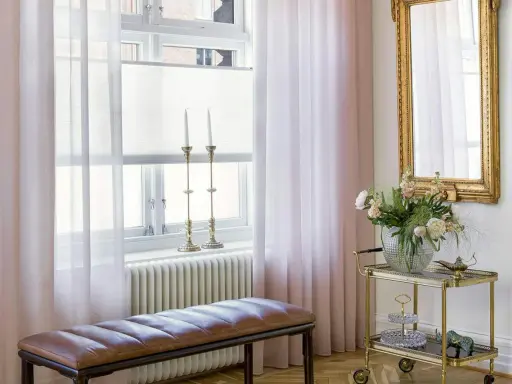 Ljusrosa gardiner längd från golvet ramar in ett fönster med ljusinsläpp. En brun läderbänk står framför ett element.