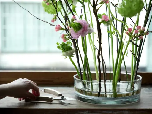 En hand förbereder vanliga snittblommor i en glasvas med en blomstergaffel. Rosa blommor och gröna detaljer skapar en vacker bukett.