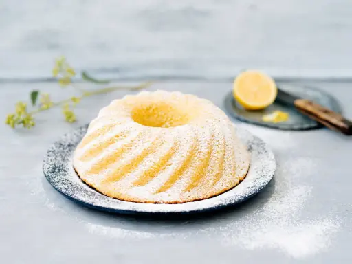 En vacker citronkaka i långpanna, pudrad med florsocker, står på ett fat. En halv citron och en kniv syns i bakgrunden.