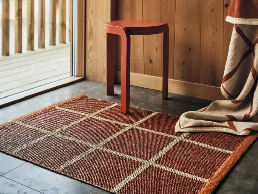 En rödbrun Pappelina Leksand-matta med rutmönster, en orange pall och en filt med randigt mönster i en mysig miljö.