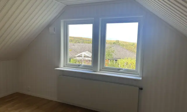 Vitt snedtak med panel och ett fönster med utsikt över träd. Gardiner saknas, men ett element finns under fönstret.