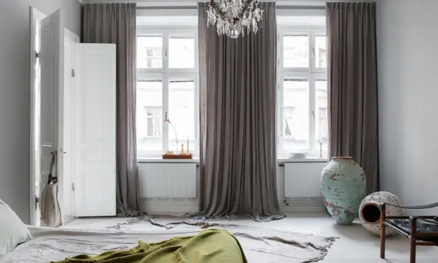 Sovrum med ljusa mörkläggningsgardiner som ramar in fönstren. En takkrona i kristall hänger ovanför.