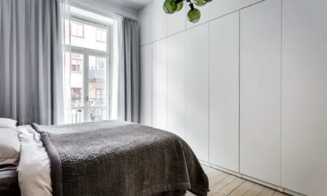 Sovrum med en stor, vit garderob med draperi. En säng med mörkt överkast står framför ett fönster med ljusgrå gardiner.