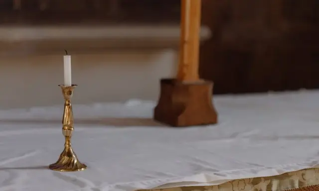 En ljusstake med ett vitt ljus står på ett vitt dukat bord. En träkorsfot skymtar i bakgrunden. Perfekt dop inspiration.