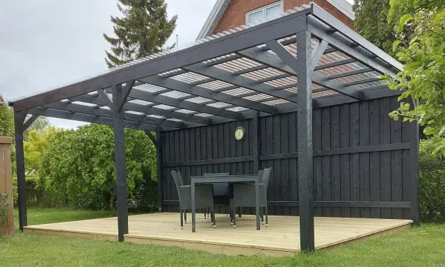 En mörk paviljong på en träaltan med bord och stolar. Perfekt för fika i trädgården.