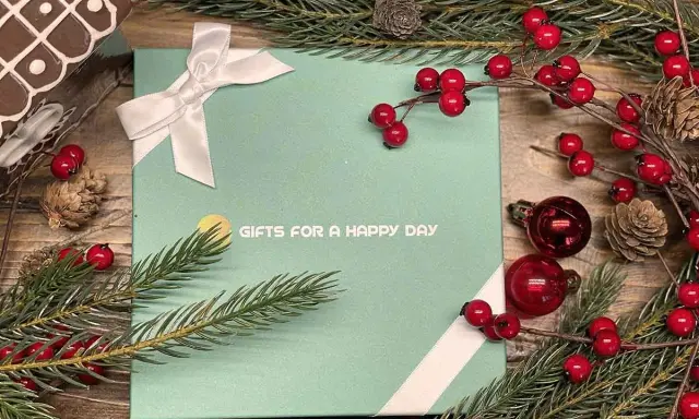En grön presentask med texten "GIFTS FOR A HAPPY DAY", dekorerad med julgranspynt, kottar och röda bär. Perfekt för julklappar och julrim.