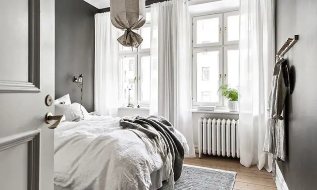 Ett sovrum med en stor säng, vita gardiner och en lång gardinstång. Mörka väggar och en dörr som står på glänt.